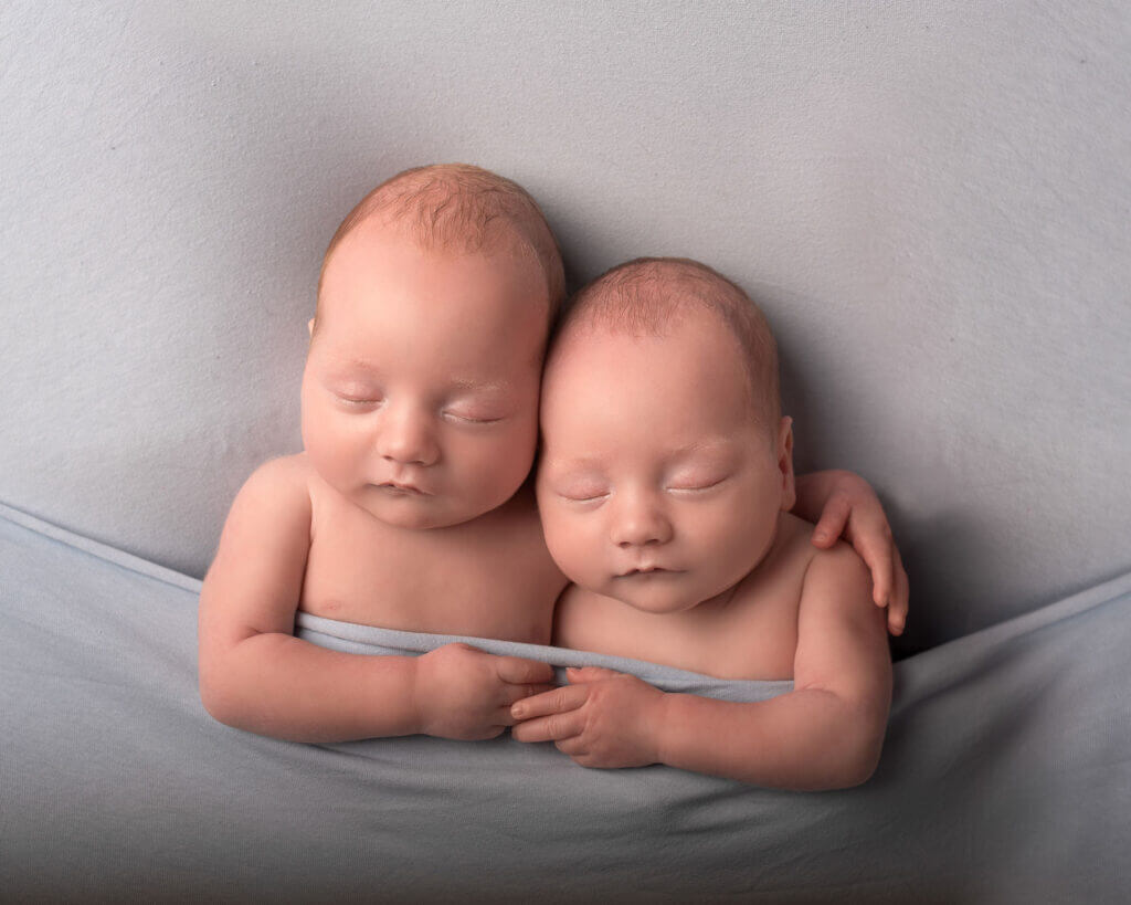 newborn baby boy twins 6 weeks old photographed in the studio lying beside each other with one twins arm around the others shoulder and holding hands tilted towards one another tucked in a pastel blue background photographed in the studio by Newborn Baby Photographer Belfast