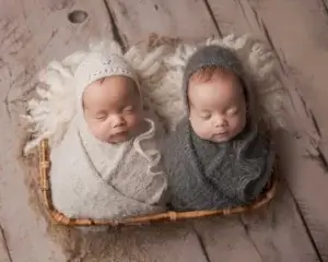 Charming image of twin baby boys peacefully sleeping side by side in a bamboo basket, one wrapped in a fluffy grey wrap and bonnet while the other is swaddled in a cream fluffy grey wrap and bonnet. Set against a clean white wooden flooring, this adorable scene captures the essence of a professional newborn photoshoot session in Belfast, showcasing the beauty of twin newborns in a serene and timeless setting
