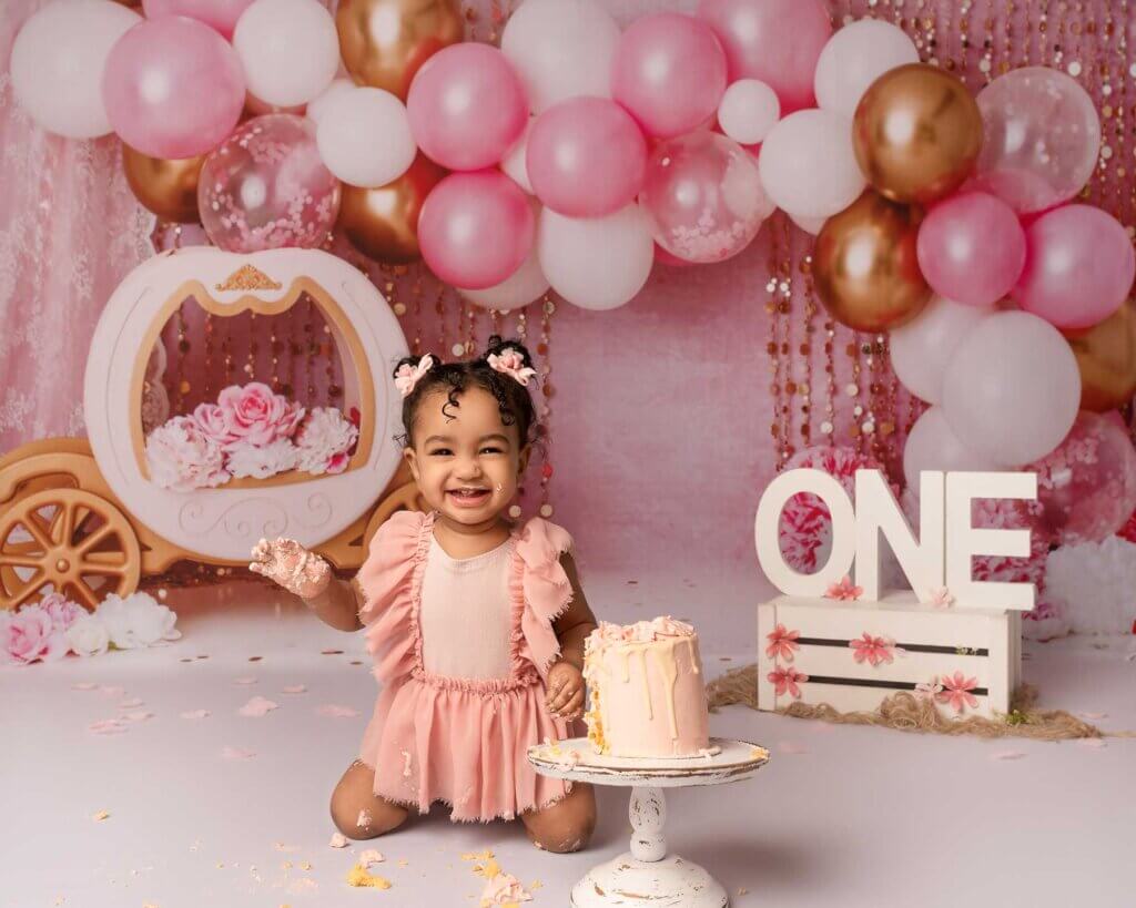 baby girl on her knees in front of pink balloons and beside a miniature cake at her Cake Smash Belfast photoshoot
