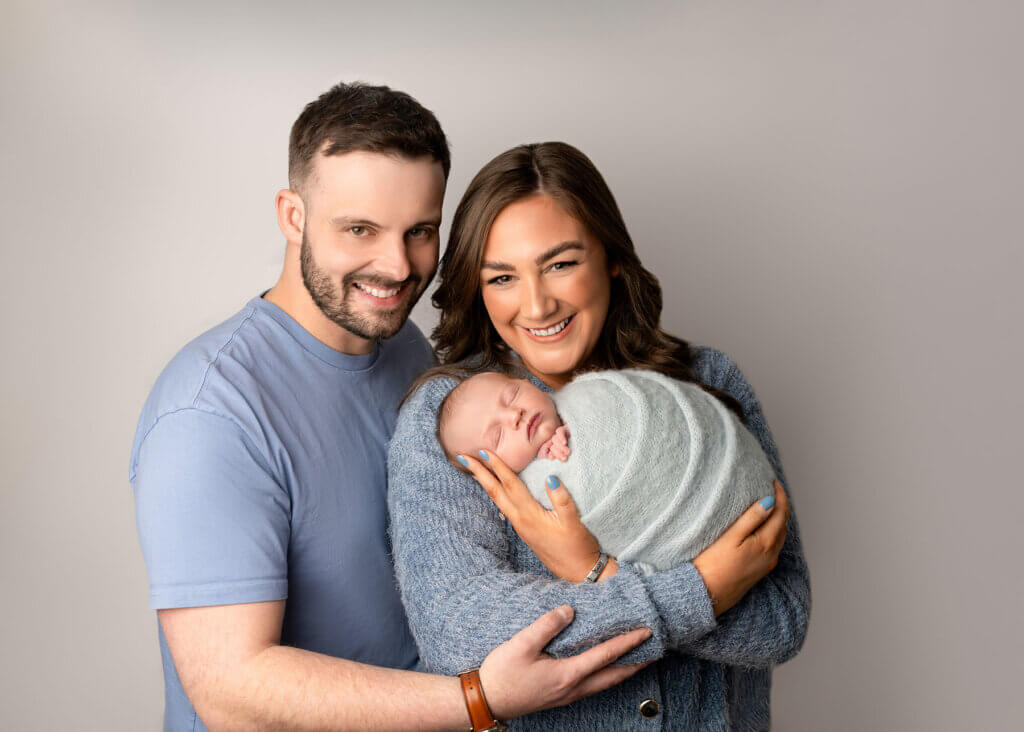 baby boy in his mummy and daddy's arms whoa re smiling at the camera by the best newborn photographer in Belfast